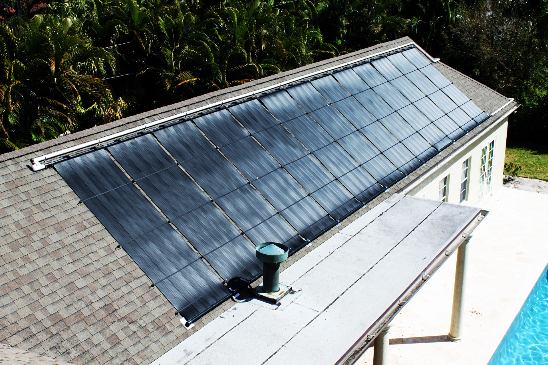 Celestial – Solar Thermal Panels Celestial Solar & Water Systems installation featuring blue-gray rectangular solar thermal panels mounted on a sloped roof beside lush tropical trees, showcasing energy efficient heating