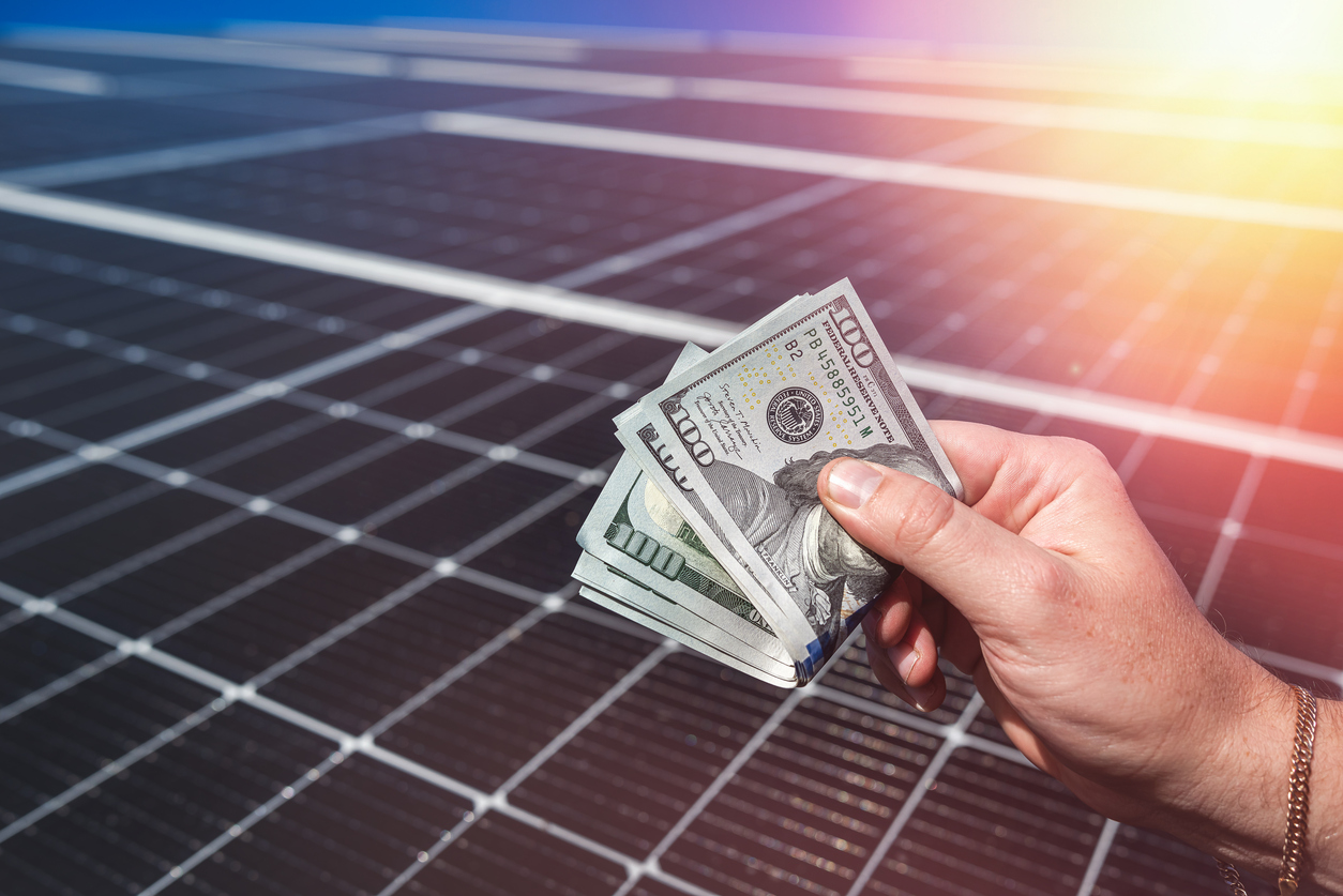 Saving money with solar energy and solar panels. Hand with dollars in front of panels. Celestial Solar & Water Systems, Inc. hand holding cash in front of solar panels, signaling solar savings and predictable energy costs with solar installation.
