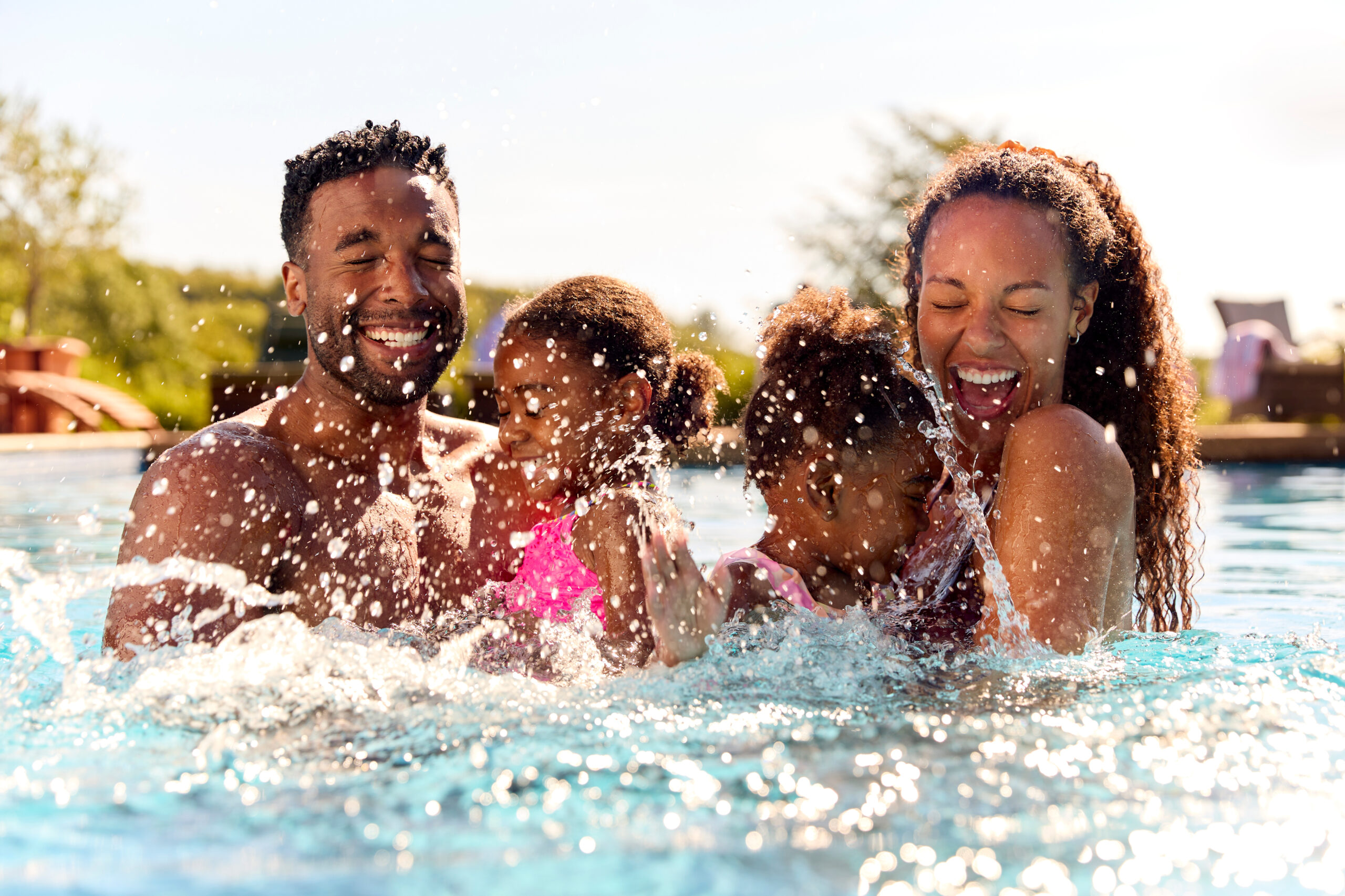 Cheerful family splashes in a sunlit outdoor pool, water droplets sparkling on blue water with a lush backyard backdrop; signals Celestial Solar & Water Systems context.