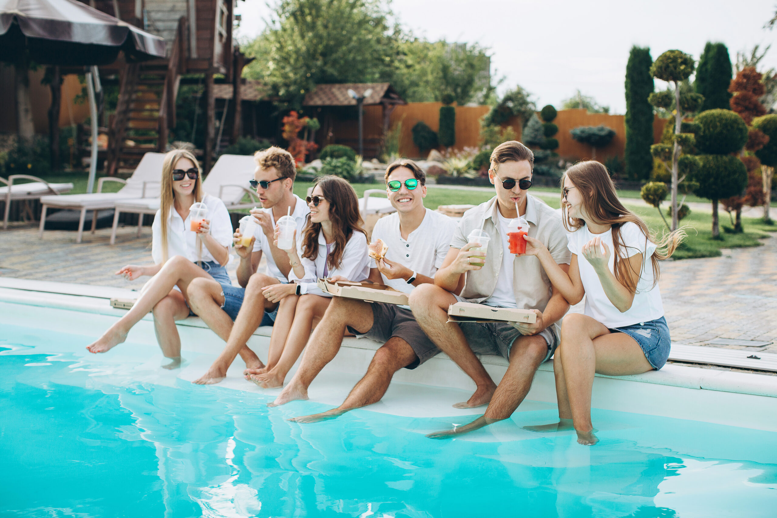 Group of young people having fun at backyard poolside party, enjoying sunny summer days outdoor, drinking cocktails and eating pizza and sushi. A sunlit outdoor pool scene with a group of friends sitting at the pool edge, sipping colorful drinks in casual white tops and shorts, by a bright blue pool and well-manicured backyard.