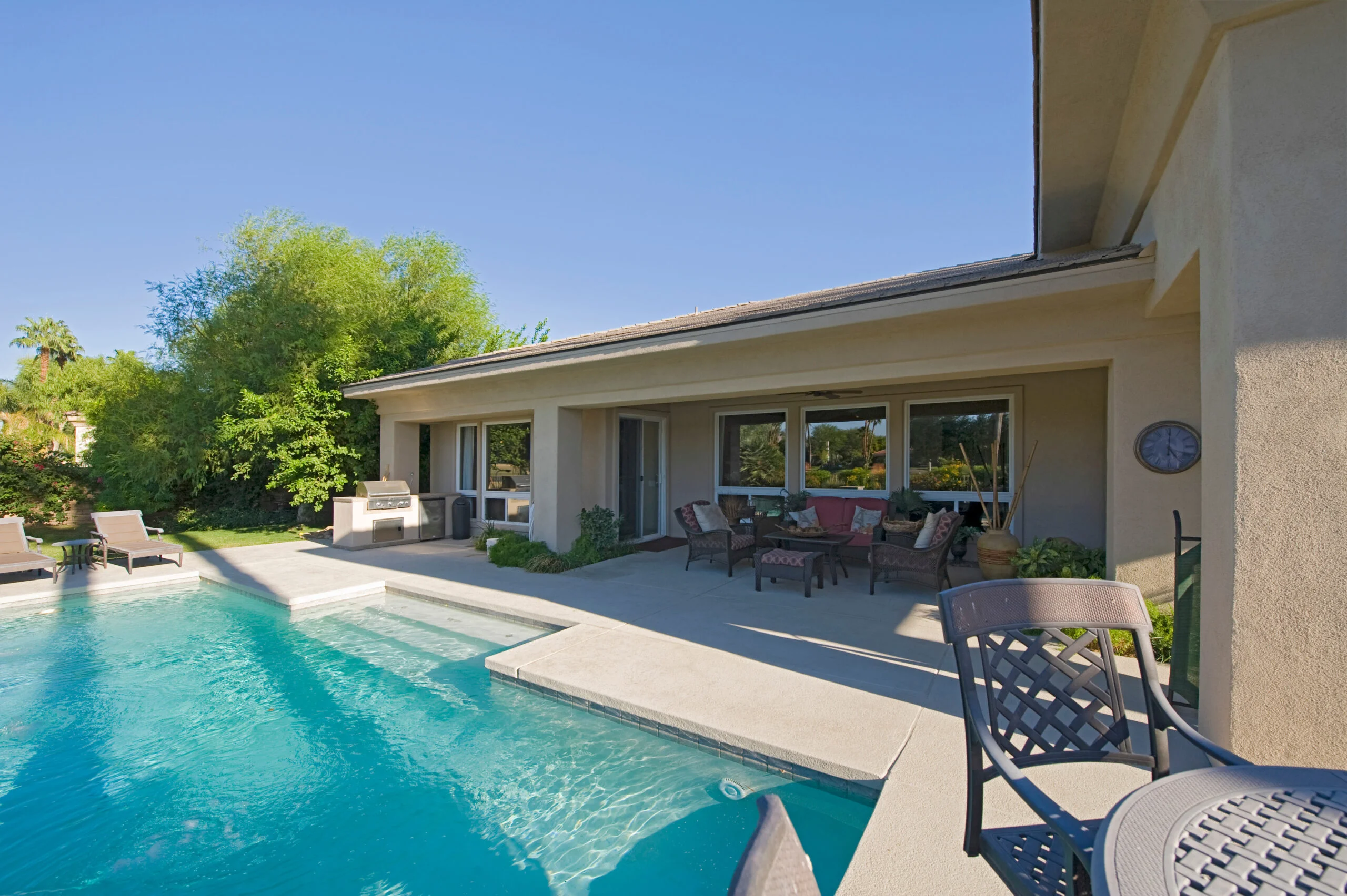 View of swimming pool in front of a modern house against sky Celestial Solar & Water Systems, Inc. desert backyard with a bright blue pool, expansive covered patio, wicker seating, potted plants, and lush greenery under a clear sky