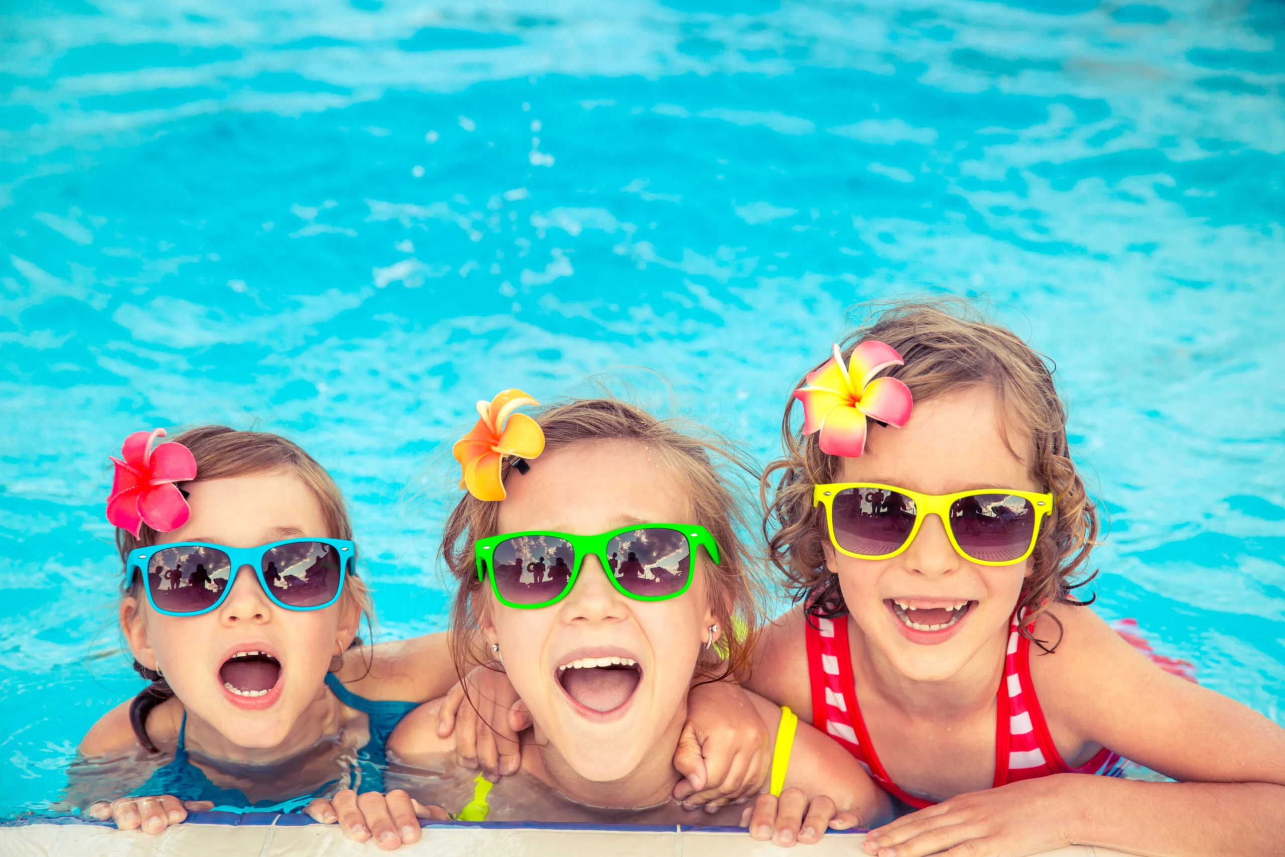Happy children in the swimming pool Spring pool season scene with three kids in a Nevada pool wearing colorful sunglasses and flower hair accents, smiling as they lean on the edge by the bright blue water.