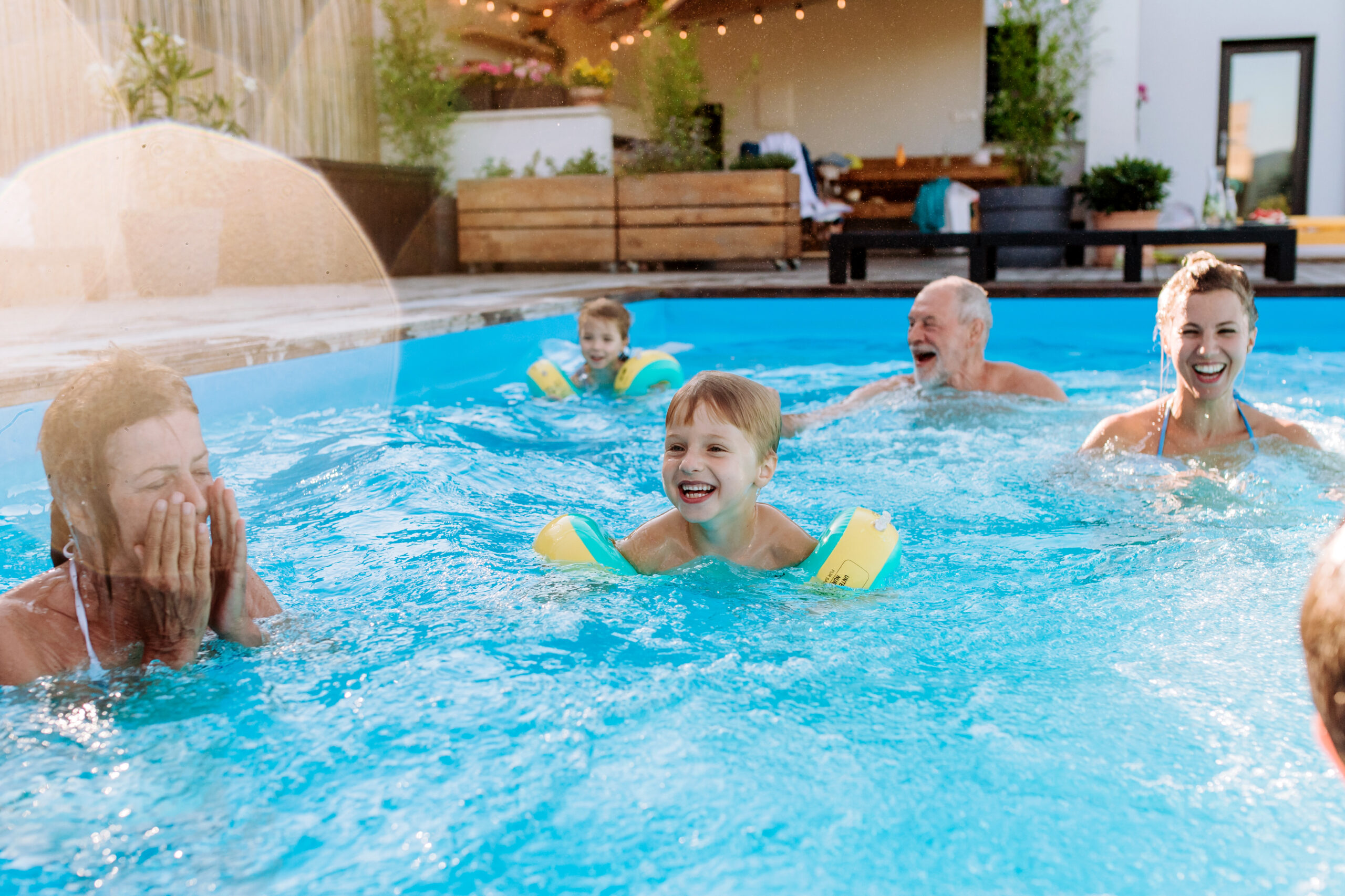 Multi generation family having fun and enjoying swimming in backyard pool during summer holiday. Bright outdoor pool scene featuring a smiling child with aqua floaties, surrounded by adults, sunlight reflecting on blue water, and a lively backyard setting