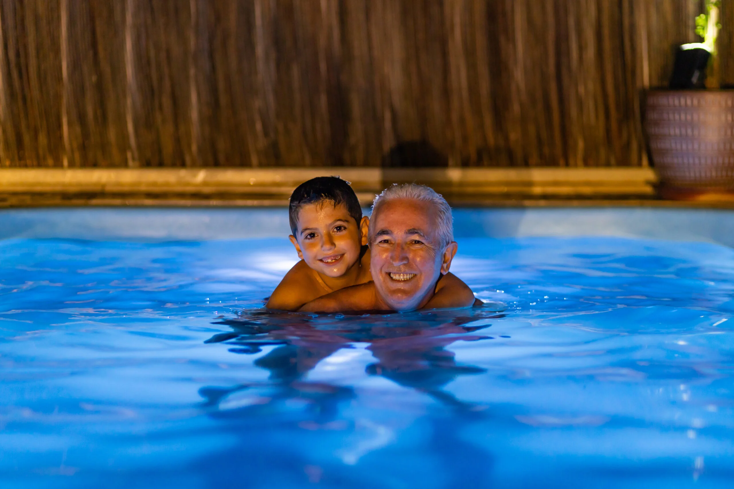 Grandfather and grandson playing in a swimming pool at night An older man and a smiling boy enjoy a nighttime swim in a glowing blue pool after sunset together.